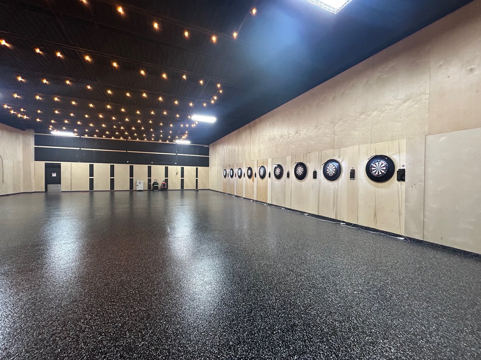 The tournament room: a long hall with dartboards down one wall, string lights overhead and a speckled epoxy floor.